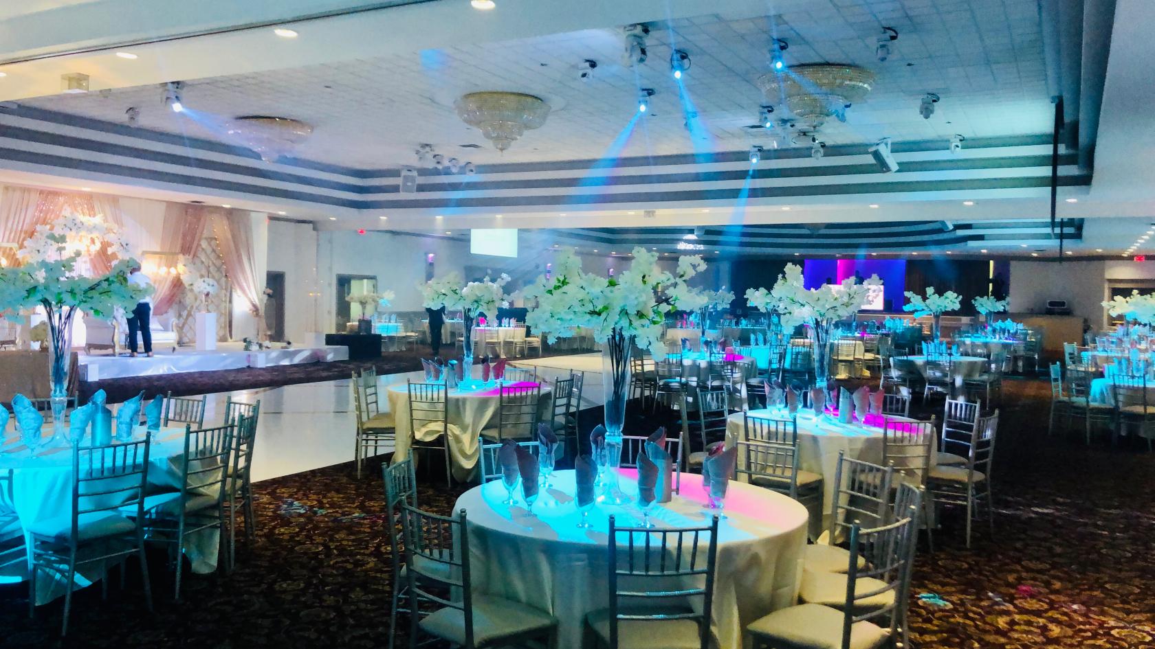 A banqueting hall with round tables, silver chairs, tall floral centerpieces, and stage lighting at Speranza Banquet Hall.
