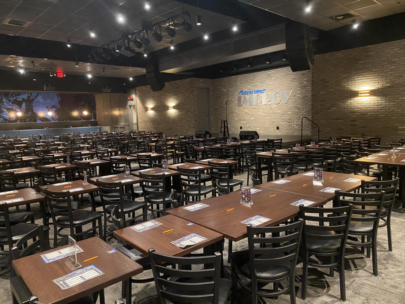 A theatre with rows of wooden tables and chairs, exposed brick walls, and stage lighting at Dania Improv.