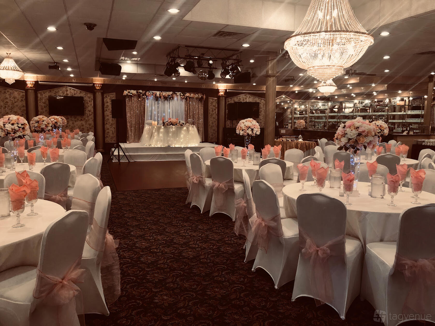 A banquet hall with round tables draped in white linens, pink chair sashes, floral centerpieces, and chandeliers at Bombay Banquet Hall.