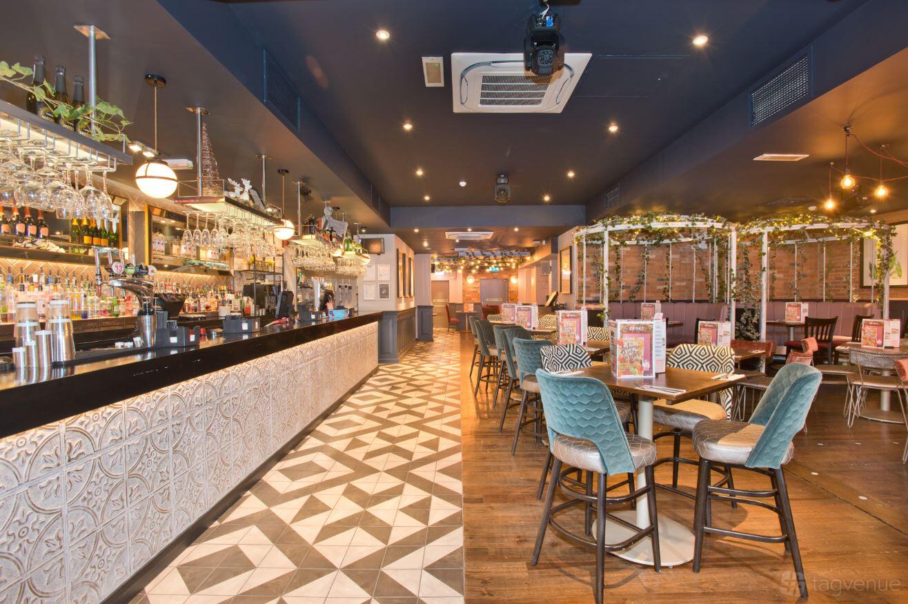 A cocktail bar with a patterned tile floor, long bar counter, hanging glassware, and upholstered chairs at Slug & Lettuce, Colchester.