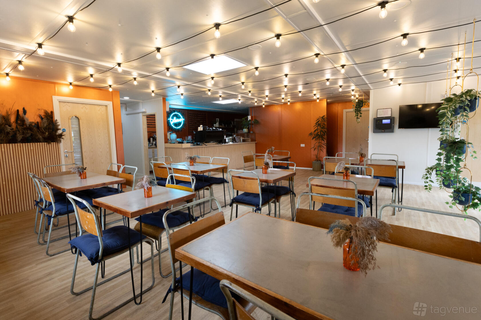 A cafe with string lights, wooden tables, metal chairs with blue cushions, and dried flower centerpieces at Firs Farm Community Hub.