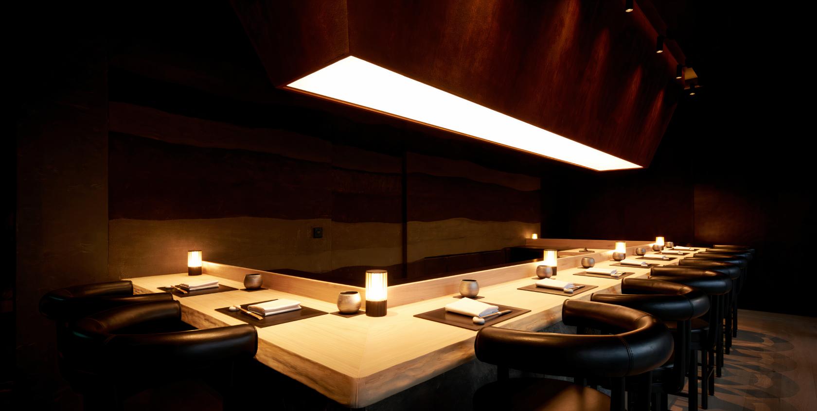 A restaurant with a light wood sushi counter, black leather stools, and soft pendant lighting at Shoukouwa Shinjidai.
