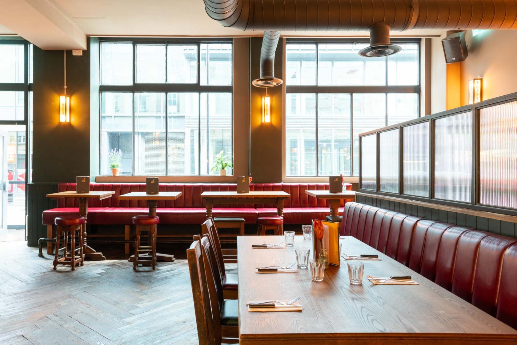 An event space in a bar with red leather banquette seating, large windows, and wooden tables at The Fountain & Ink.