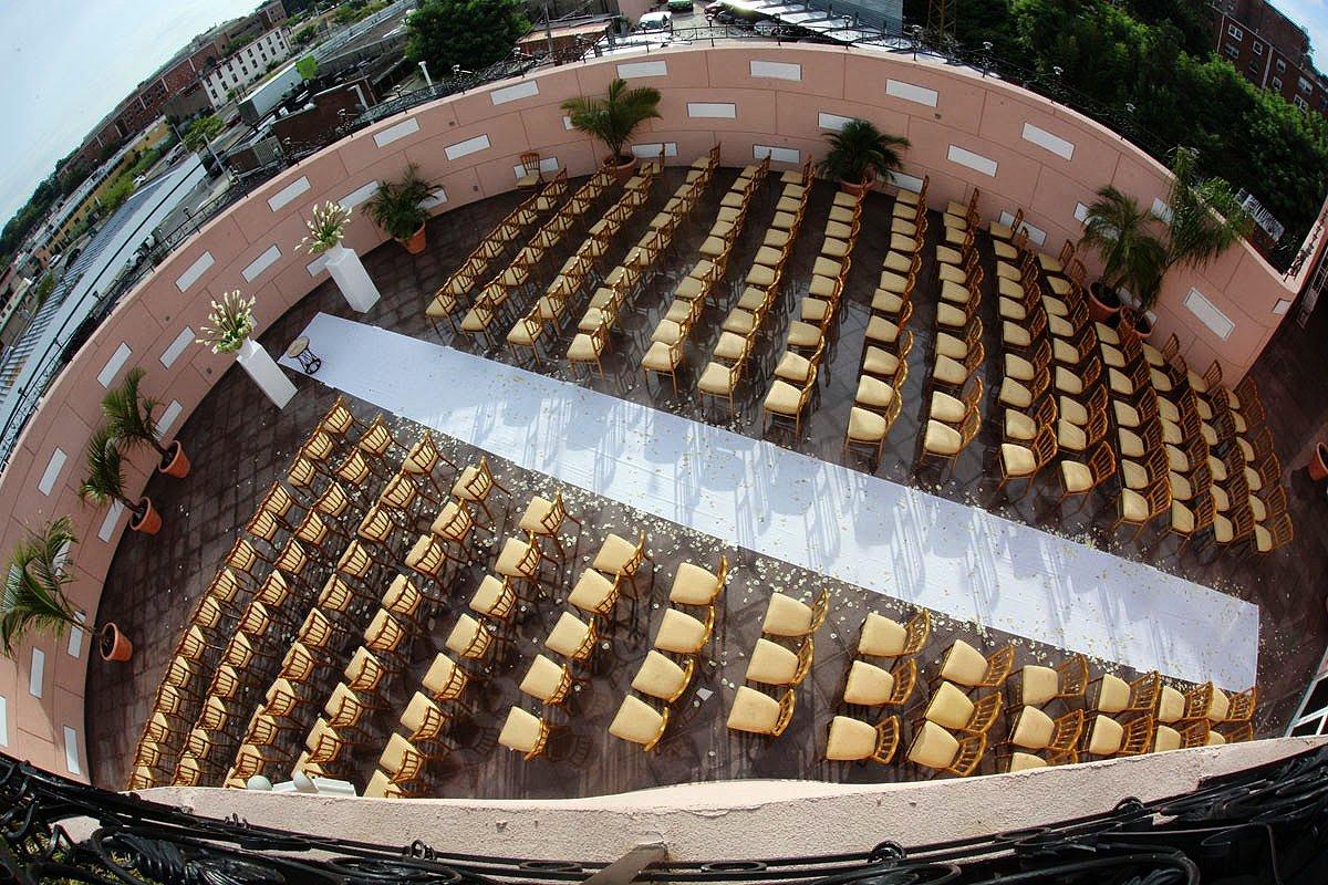 A rooftop event space with rows of gold chairs, white aisle runner, and potted palm trees at Da Mikele Illagio.