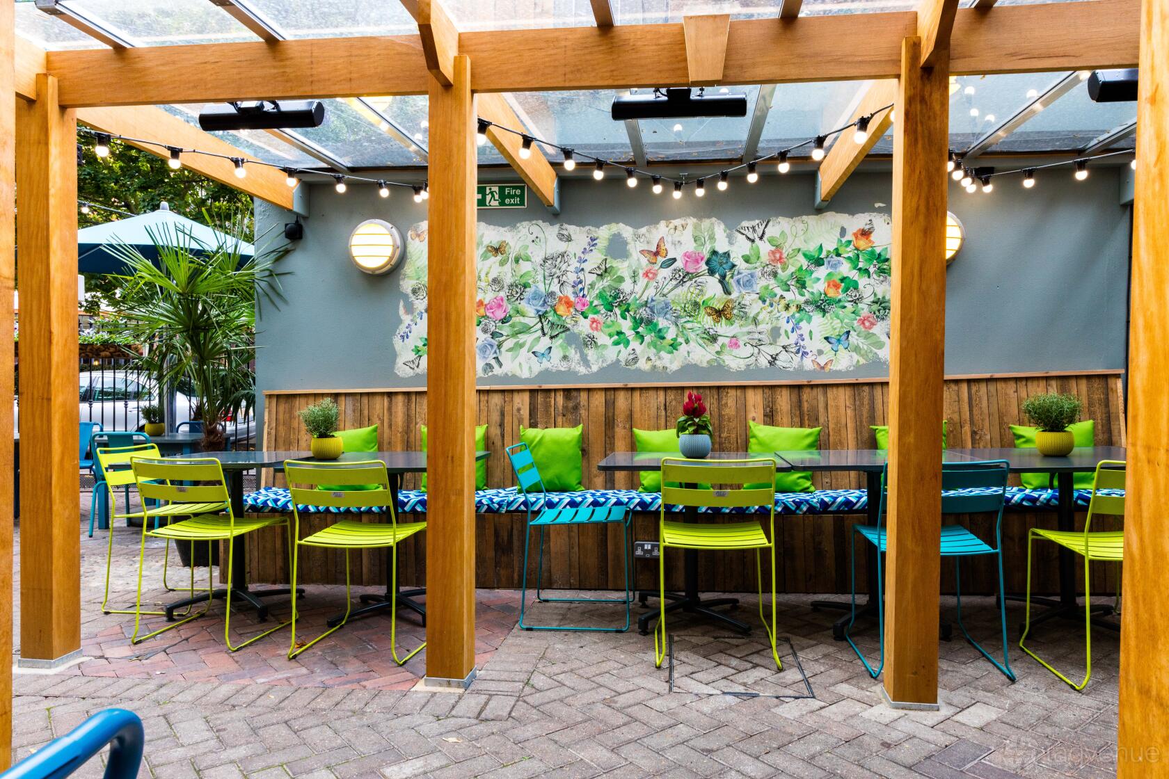 An outdoor space with a covered pergola, string lights, and colorful chairs at Brook Green.