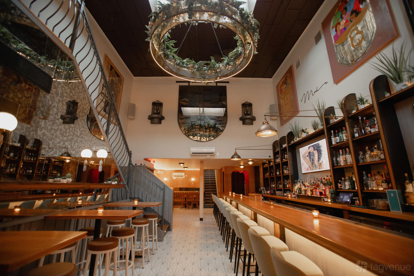 A bar with a long wooden counter, high stools, greenery chandelier, and shelving with bottles at Madeline's.