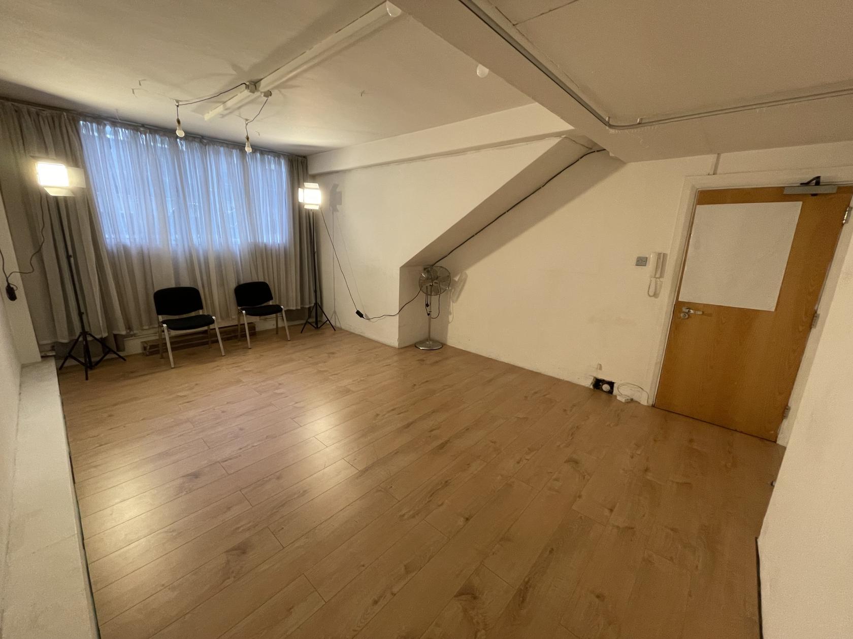 A dance studio with light wood flooring, two black chairs, standing lamps, and a curtain over large windows at Studio Movement 2.