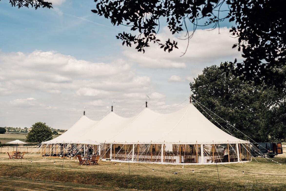 An outdoor event venue with a large white marquee tent and wooden tables on grass at Peacock and Bow.