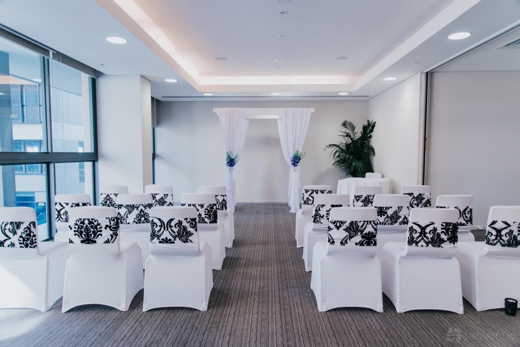 An event space with rows of white chairs with patterned covers, a sheer draped arch, and floor-to-ceiling windows at Novotel Newcastle Beach.