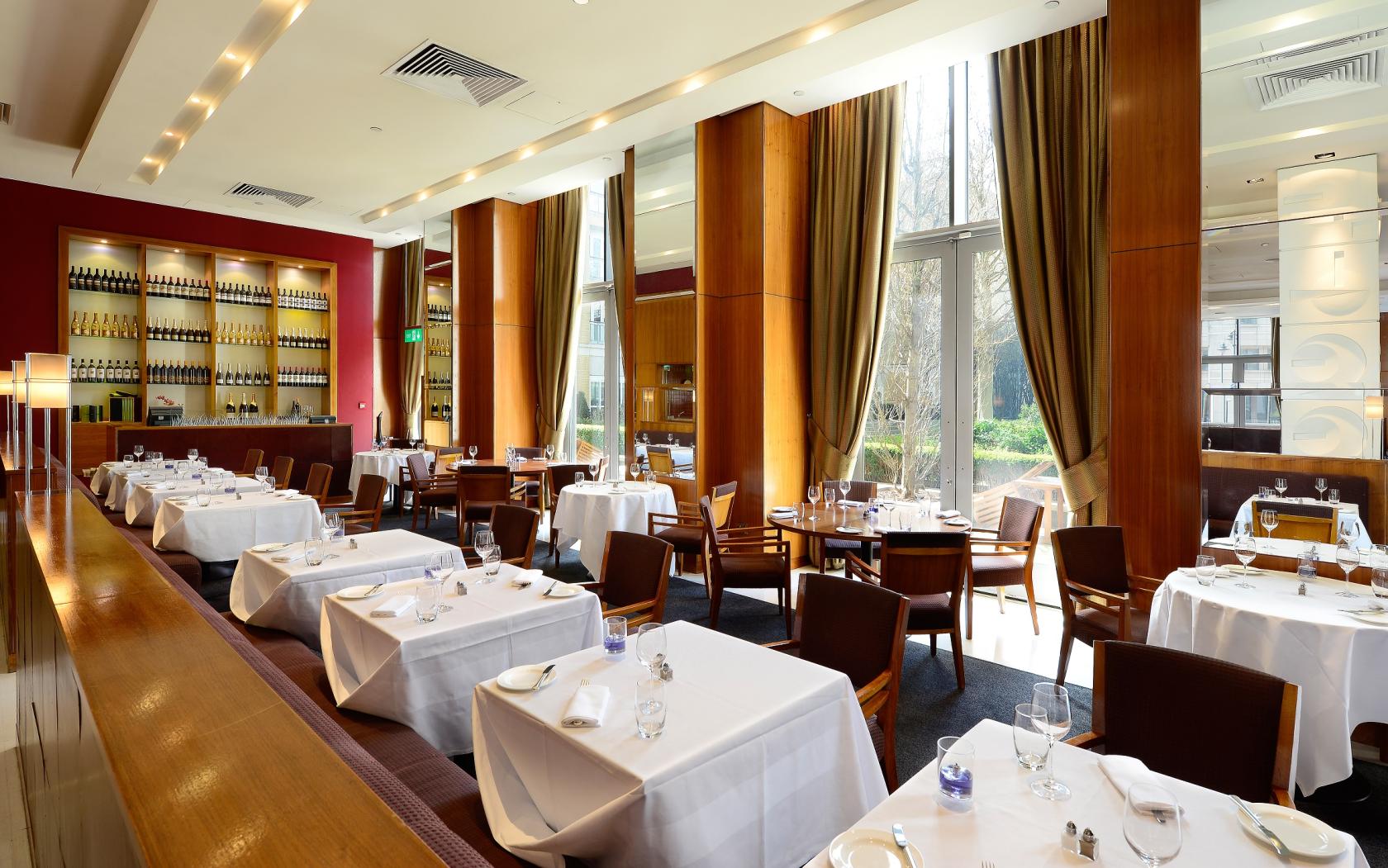 A restaurant with floor-to-ceiling windows, white tablecloths, and wooden shelving at Canary Riverside Plaza Hotel.