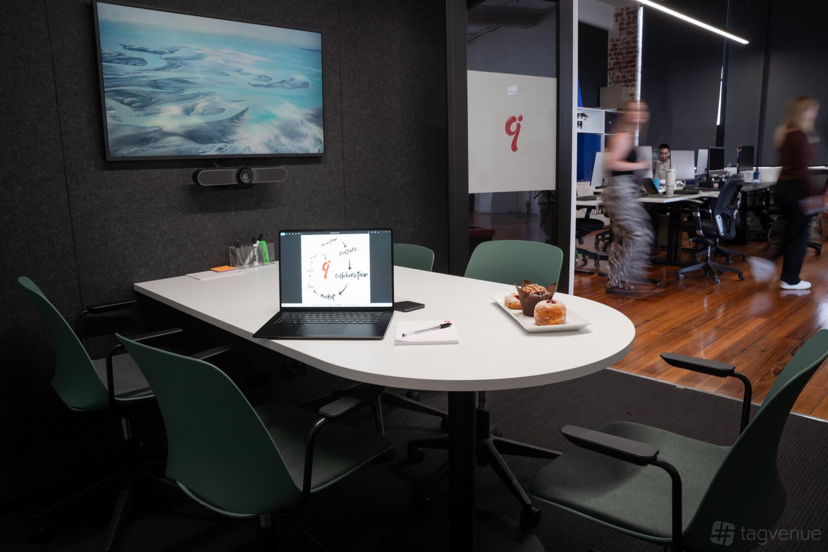 A meeting room with a wall-mounted screen, conference table, laptop, and pastries at Inspire9.
