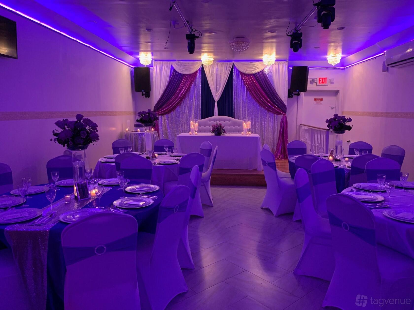 A hall with purple uplighting, round tables with white linens, and a decorated stage at La Belvedere LLC.