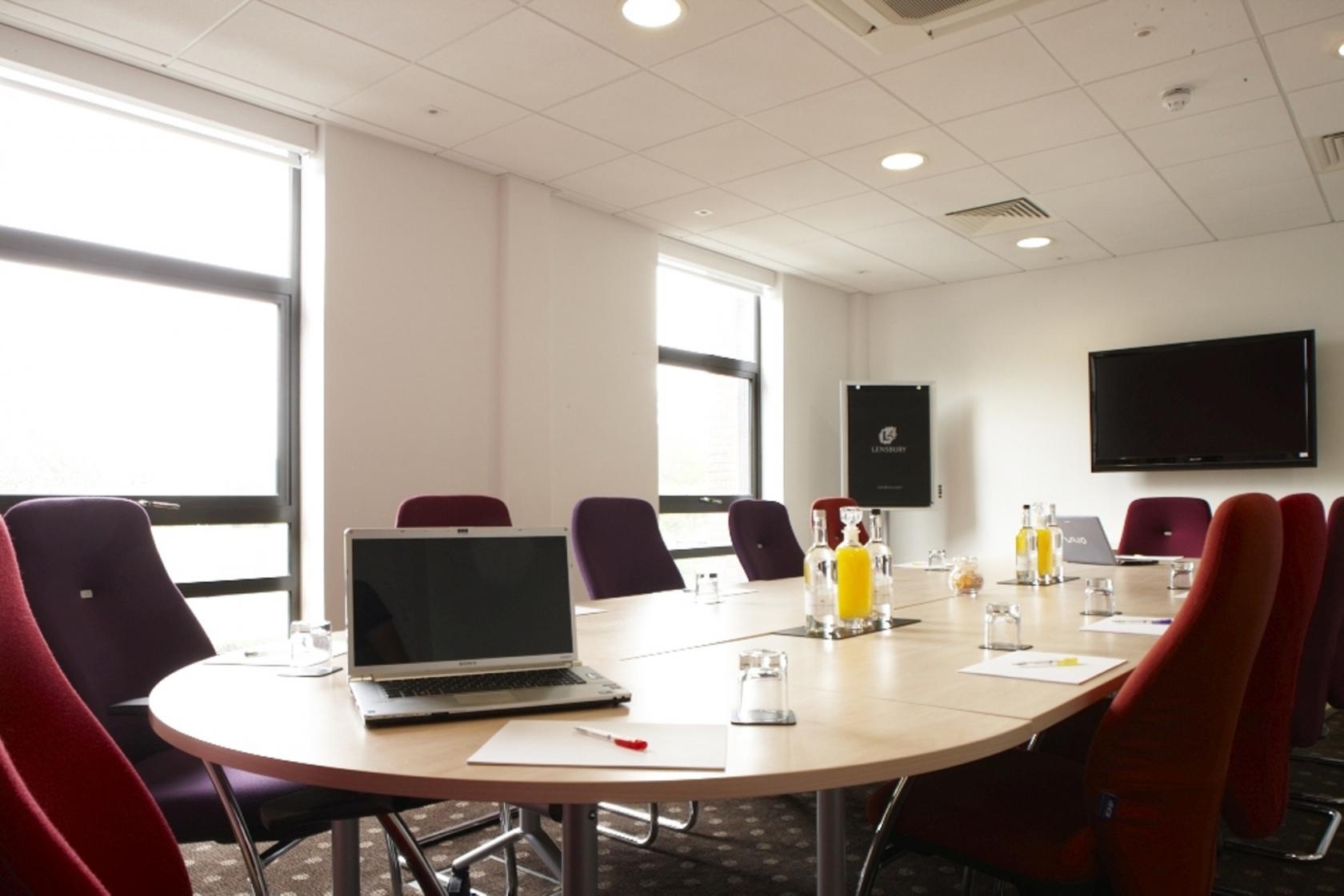 A hotel meeting room with oval table, purple chairs, large windows, and a wall-mounted screen at The Lensbury.