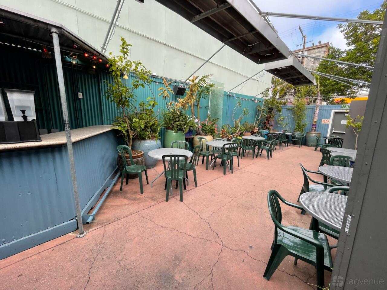 An outdoor terrace with metal tables, green plastic chairs, potted plants, and a corrugated bar at Mars Bar Restaurant.