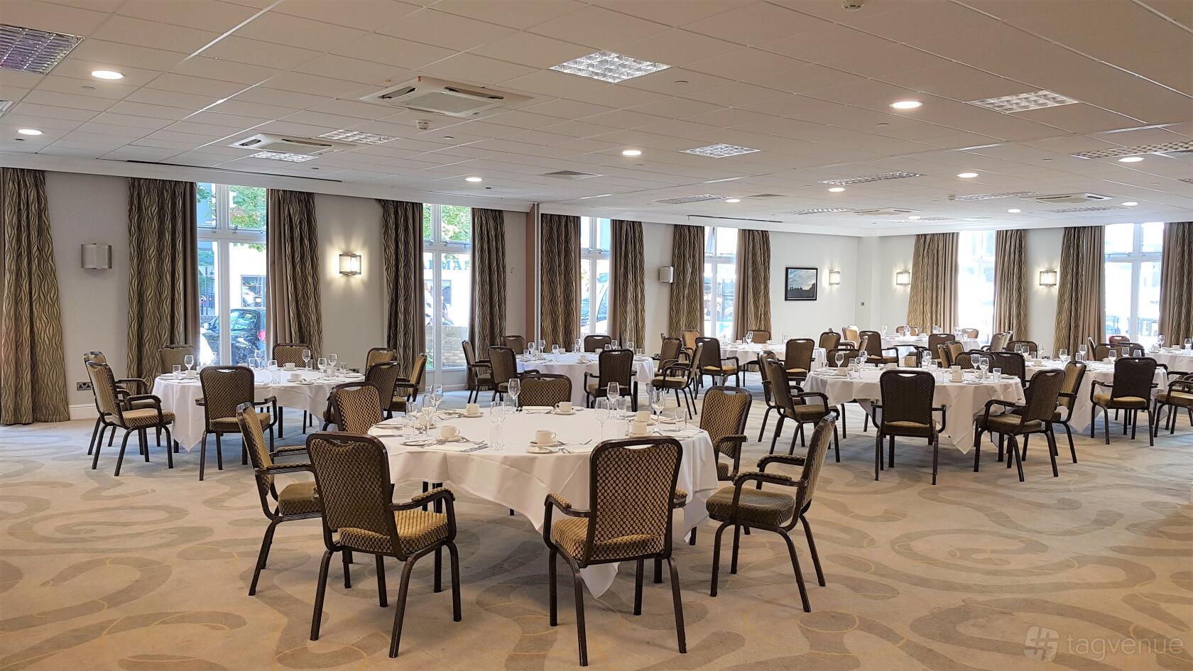 A meeting room with round tables set with white linens, banquet chairs, and large windows at Holiday Inn Kenilworth-Warwick.