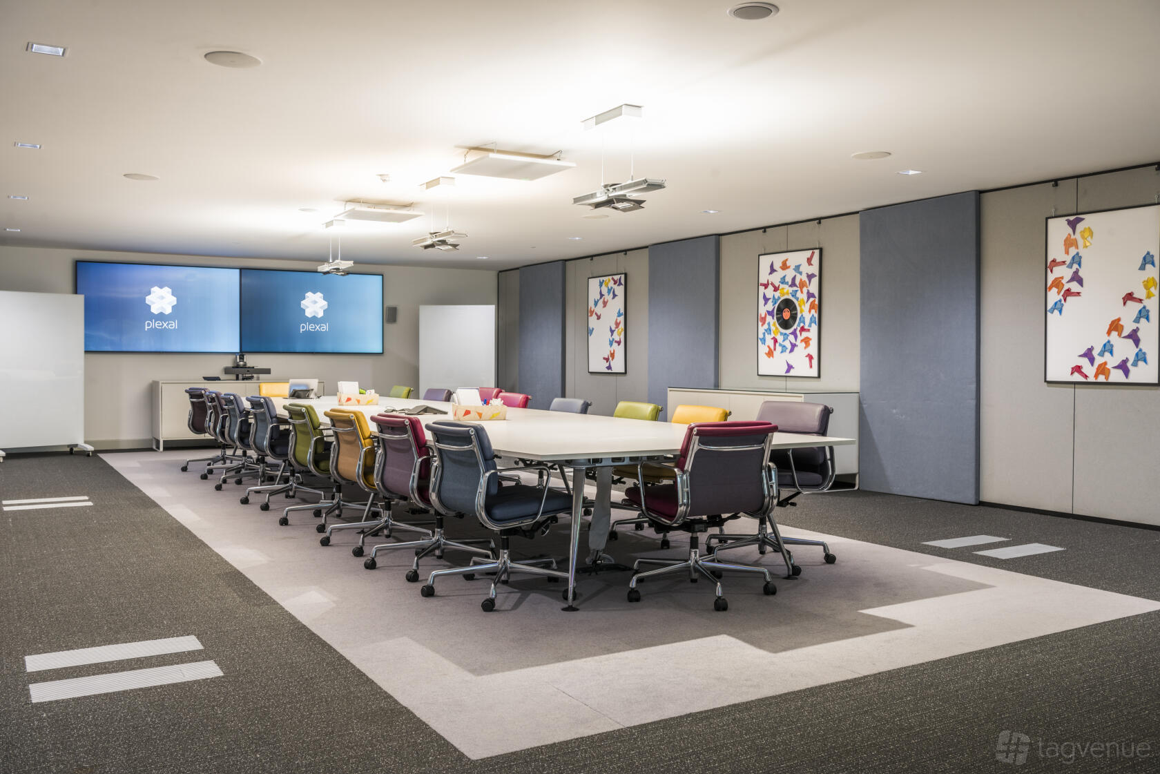 A boardroom with two large screens, colorful chairs, and framed art on the walls at PLEXAL.