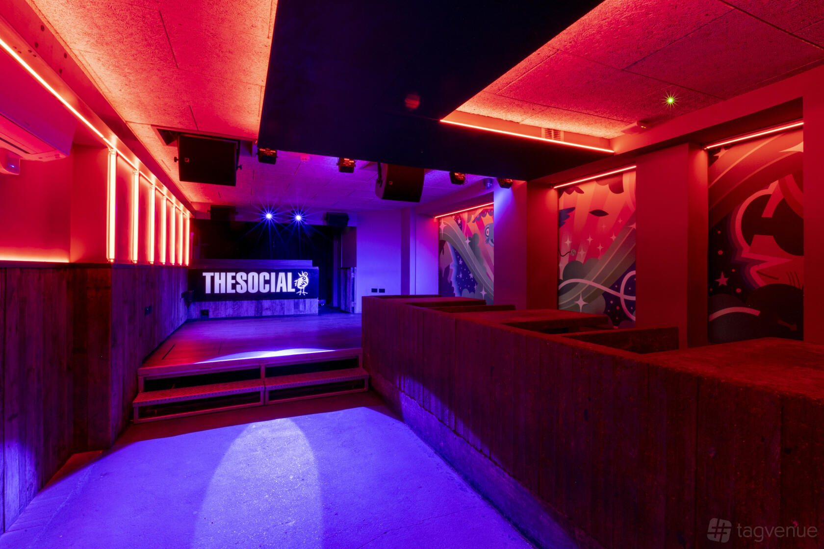 A basement bar with colorful LED lighting, a small stage, and mural-covered walls at The Social.