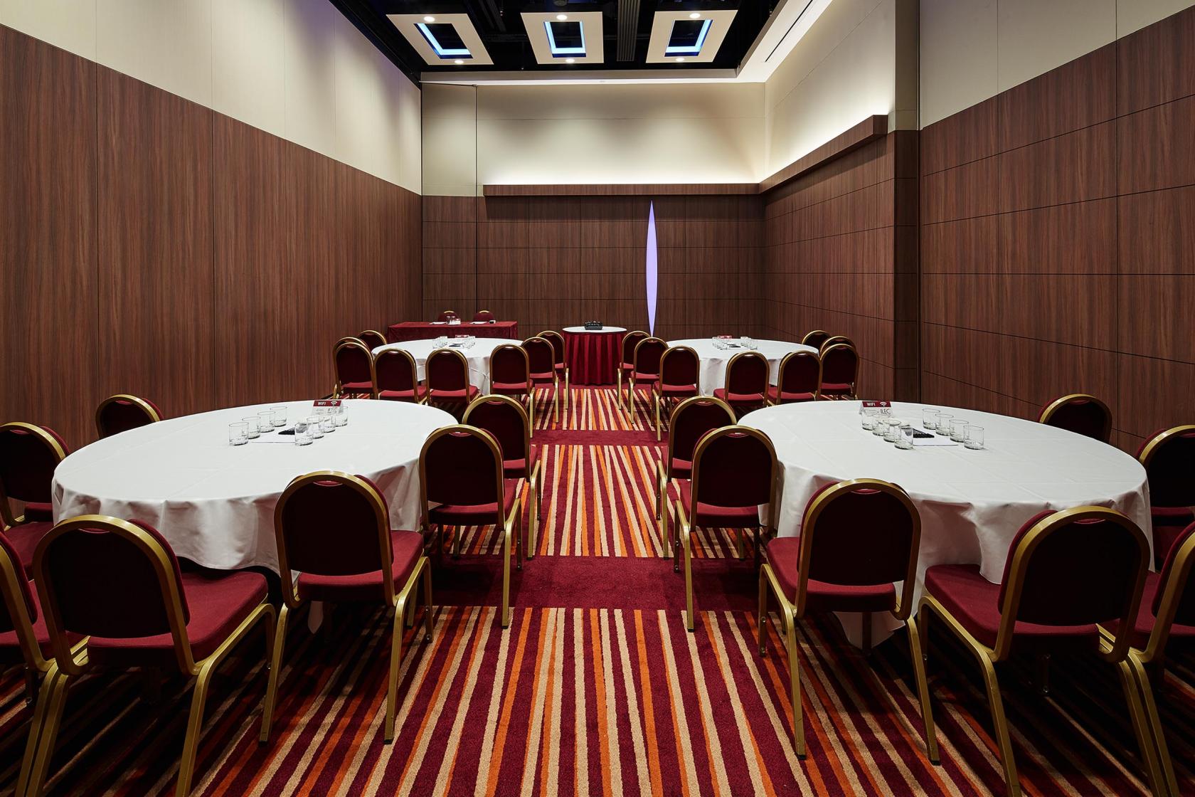A hotel meeting room with round tables covered in white linens, striped carpeting, and wood-paneled walls at Mercure London Earl's Court.