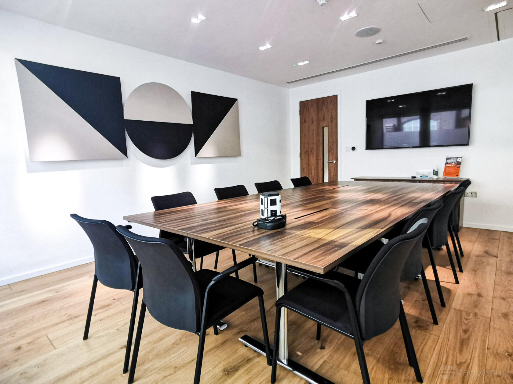 A boardroom with a long wooden table, black chairs, wall-mounted TV, and geometric art at Us&Co Stratford.