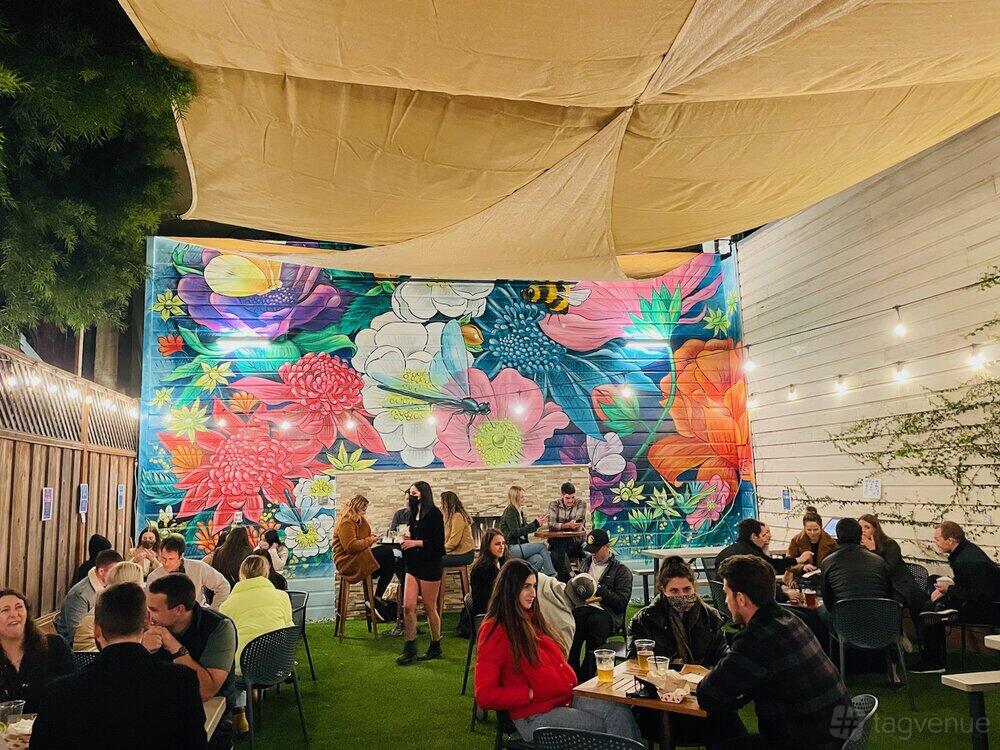 A garden at a bar with colorful floral mural, string lights, and outdoor seating on artificial grass at Campus.