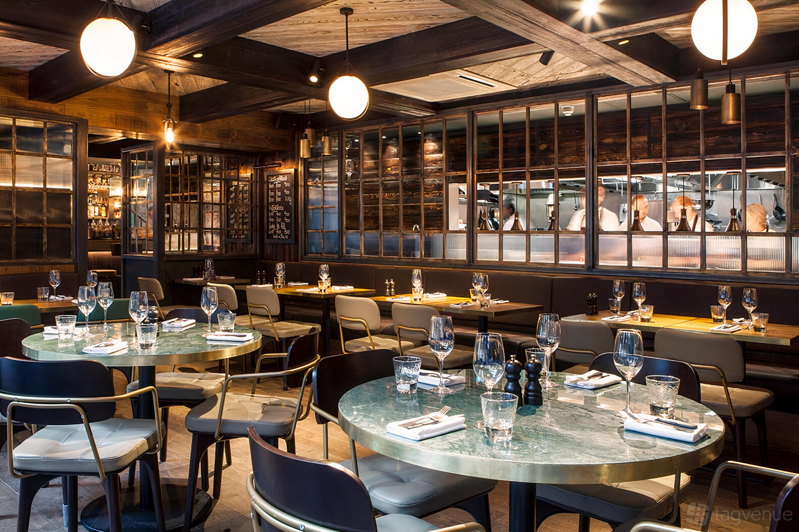 A restaurant with round marble tables, upholstered chairs, exposed wood beams, and glass partition at The Coal Shed- London.