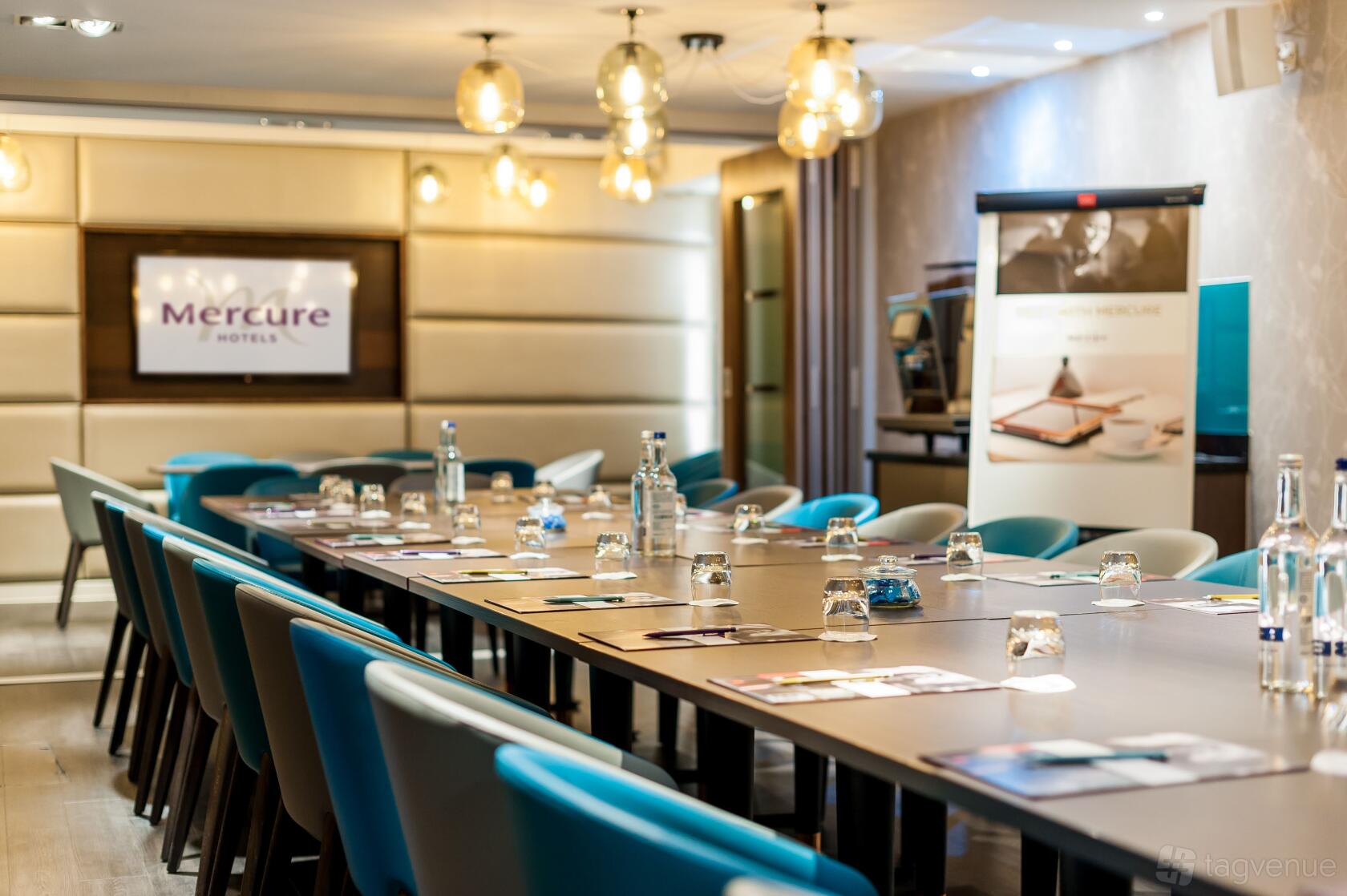 A meeting room with a long conference table, teal chairs, pendant lights, and presentation screens at Mercure London Hyde Park.