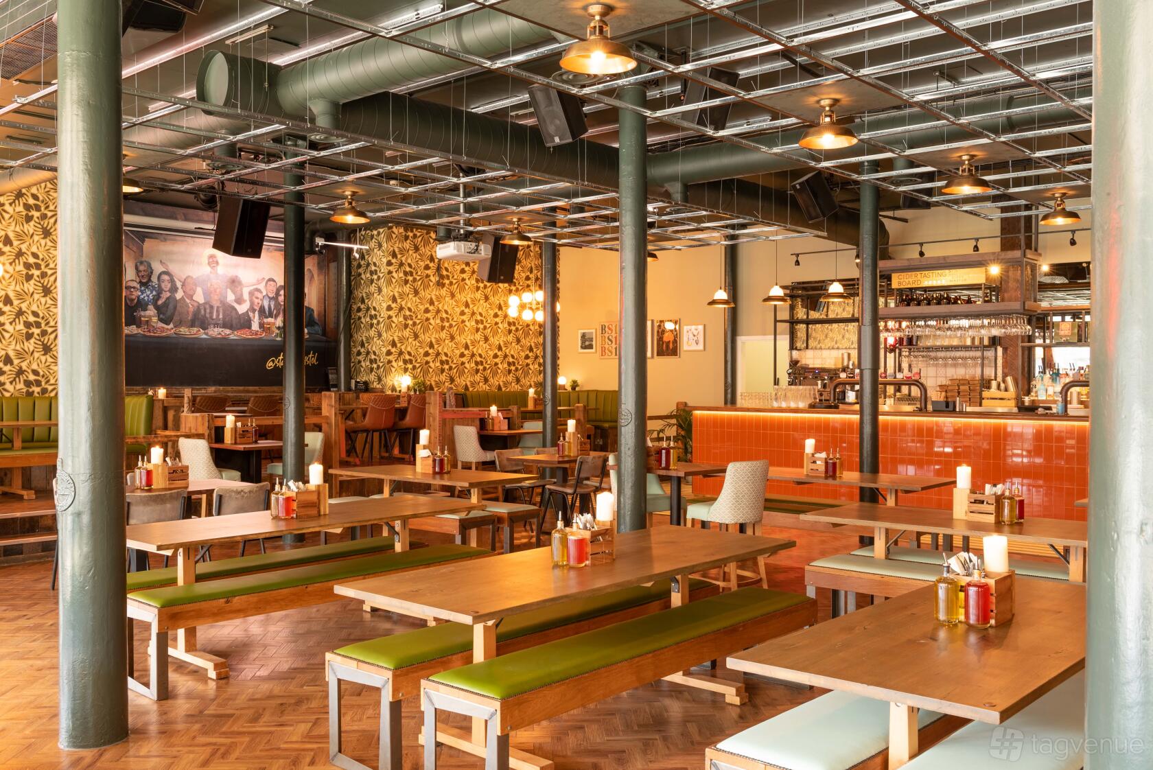 A pub with long wooden tables, green bench seating, exposed ductwork, and a mural at The Bristol Stable.