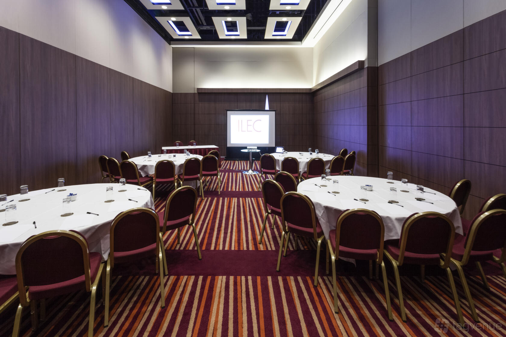 A function room with round tables, red chairs, striped carpet, and a projector screen at Mercure London Earl's Court.
