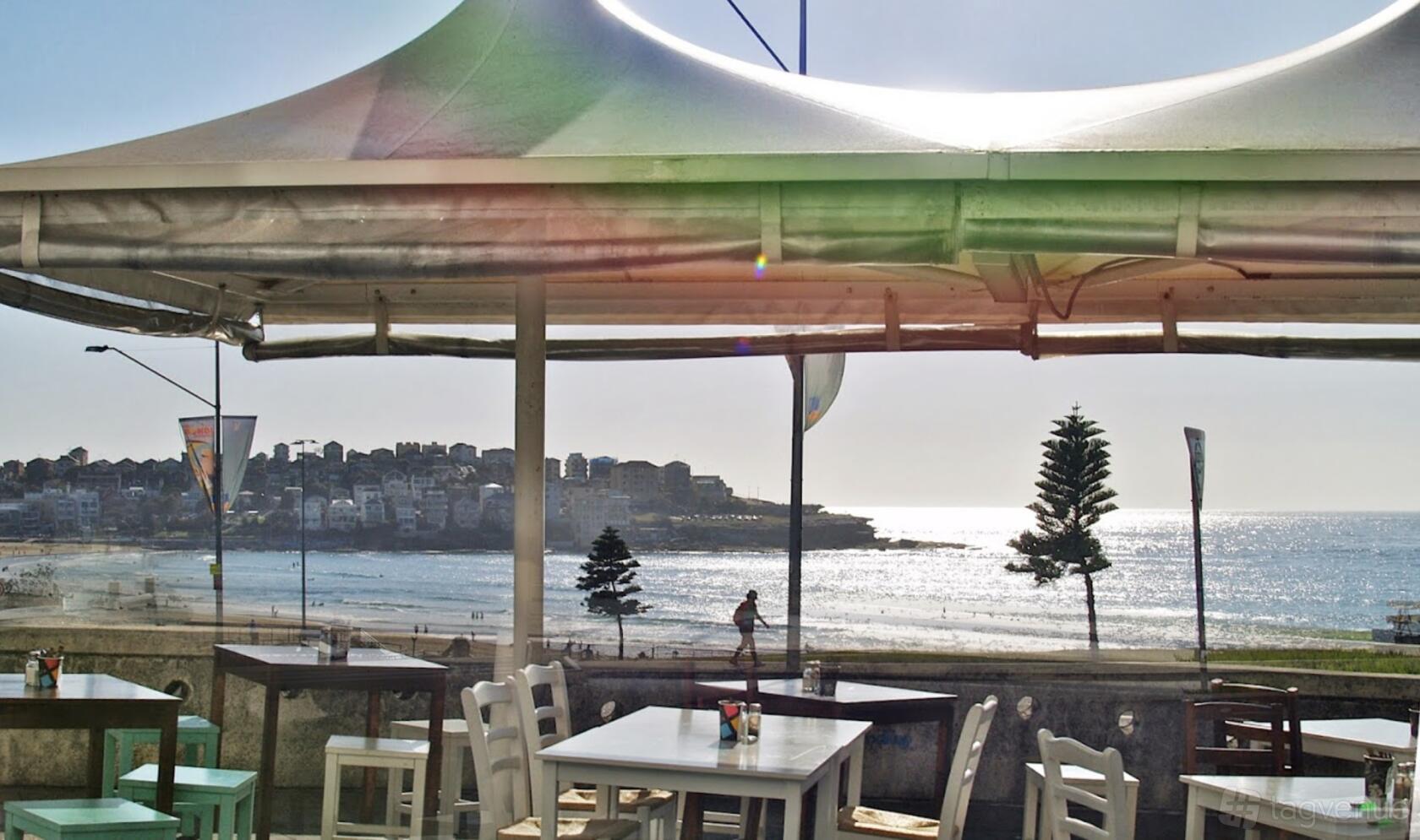 An outdoor cafe space with white tables, pastel chairs, and beach views under a canopy at Lamrock Cafe Bondi Beach.