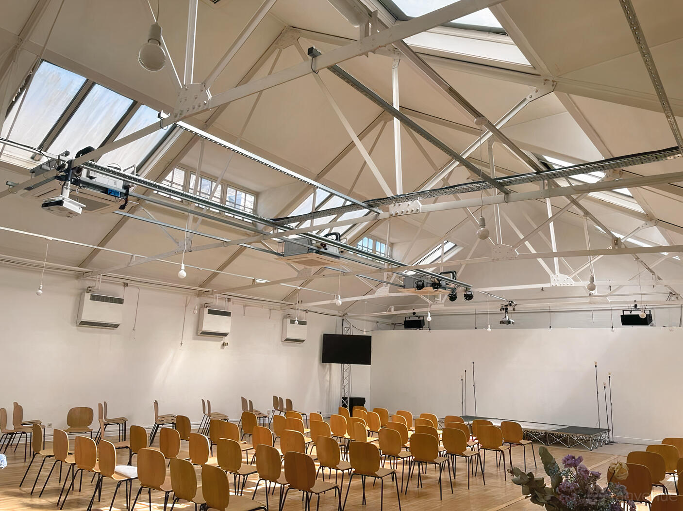 An event venue with skylights, exposed ceiling beams, and rows of wooden chairs facing a small stage at 1-3 Coborn Street.