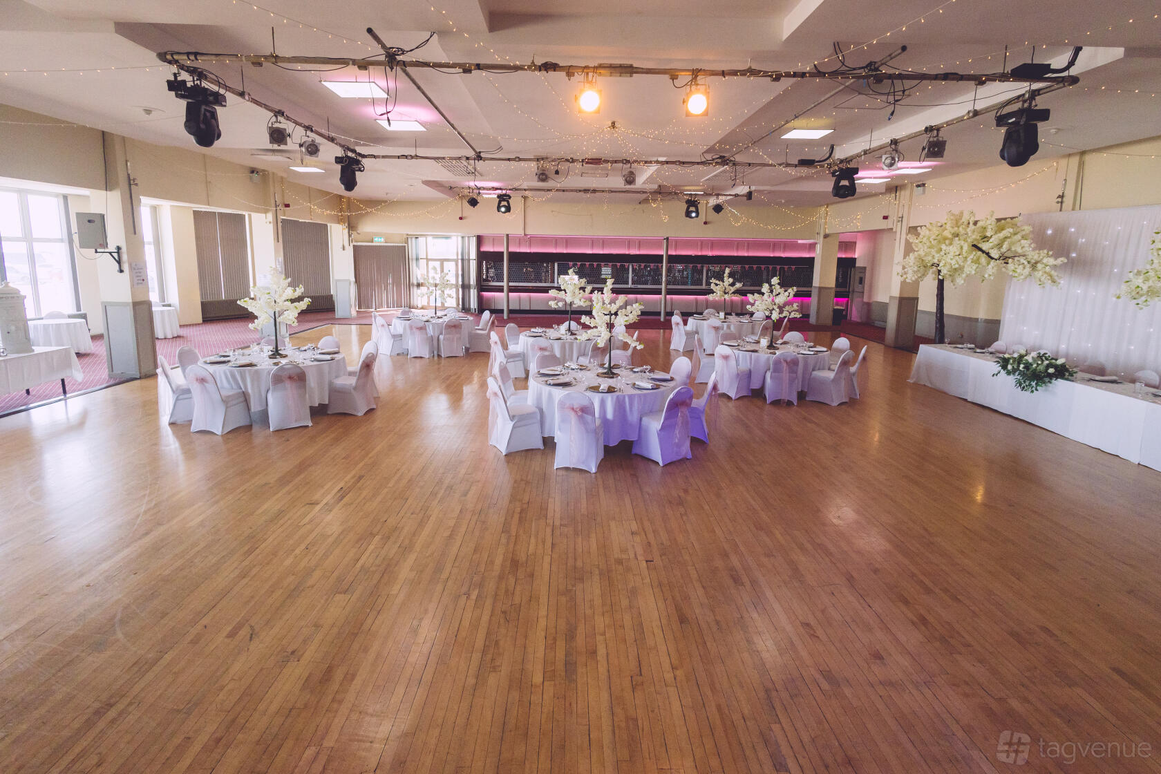 An event venue with a spacious wooden floor, round tables draped in white linens, and large windows at Weymouth Pavilion.