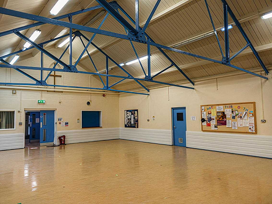 A hall with high ceilings, exposed blue steel beams, wooden floors, and noticeboards at The Newman Community Centre.