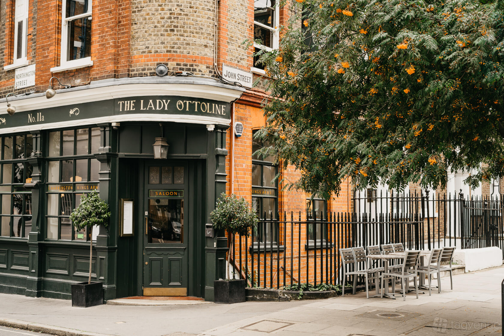 A corner pub with green exterior, large windows, brick facade, and outdoor seating under a flowering tree at The Lady Ottoline.