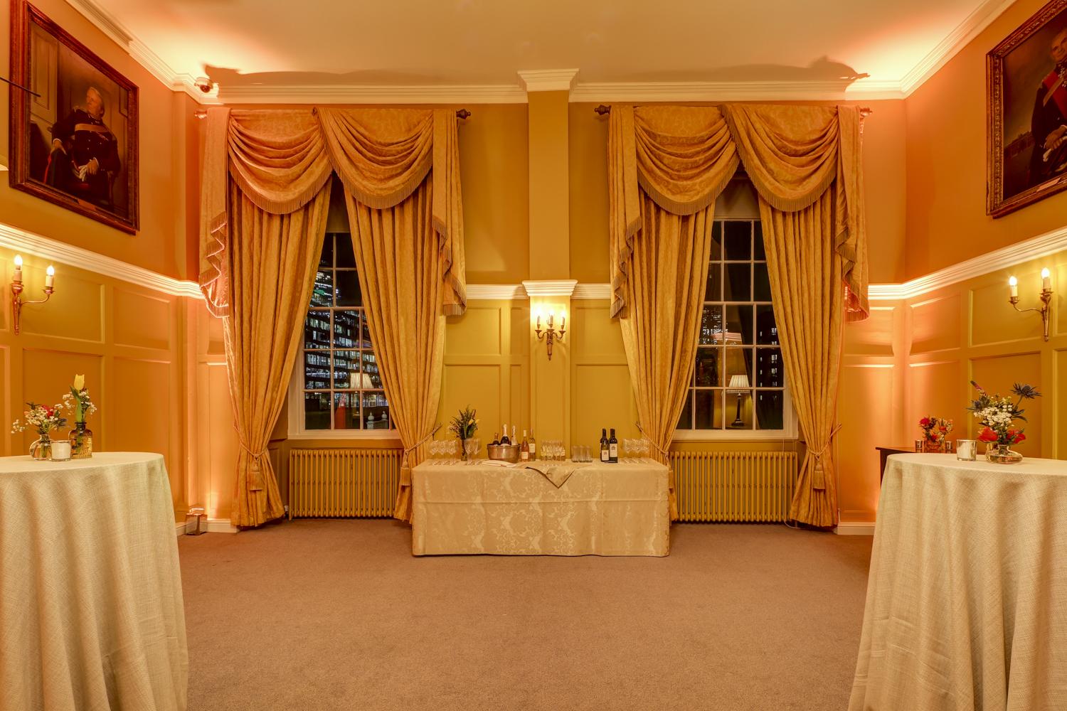 A dining room with tall windows, gold drapes, wall sconces, and round cocktail tables at The HAC.