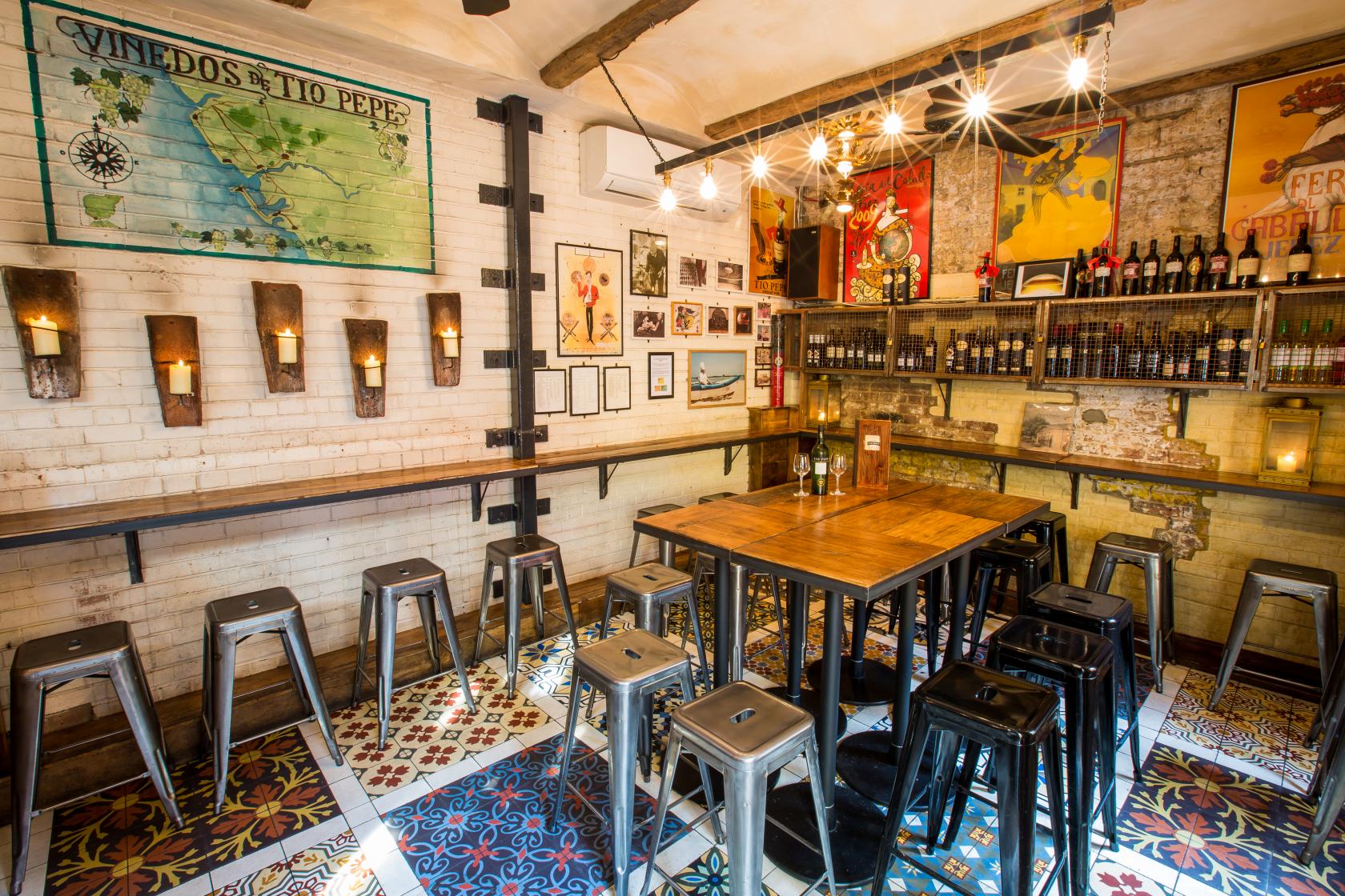 A bar with patterned tile flooring, high tables, metal stools, and wine bottles displayed on shelves at Bar Rioja.