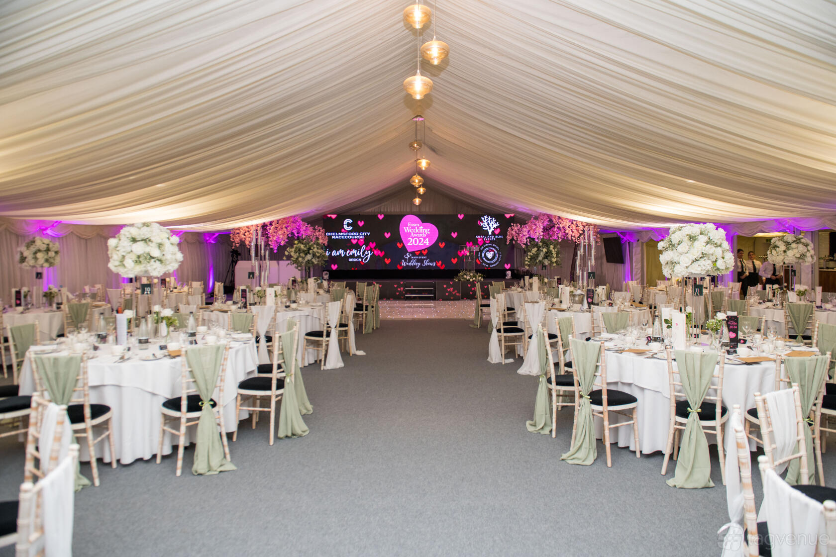 An event venue with a draped marquee ceiling, round tables with white linens, and a stage at Chelmsford City Racecourse.
