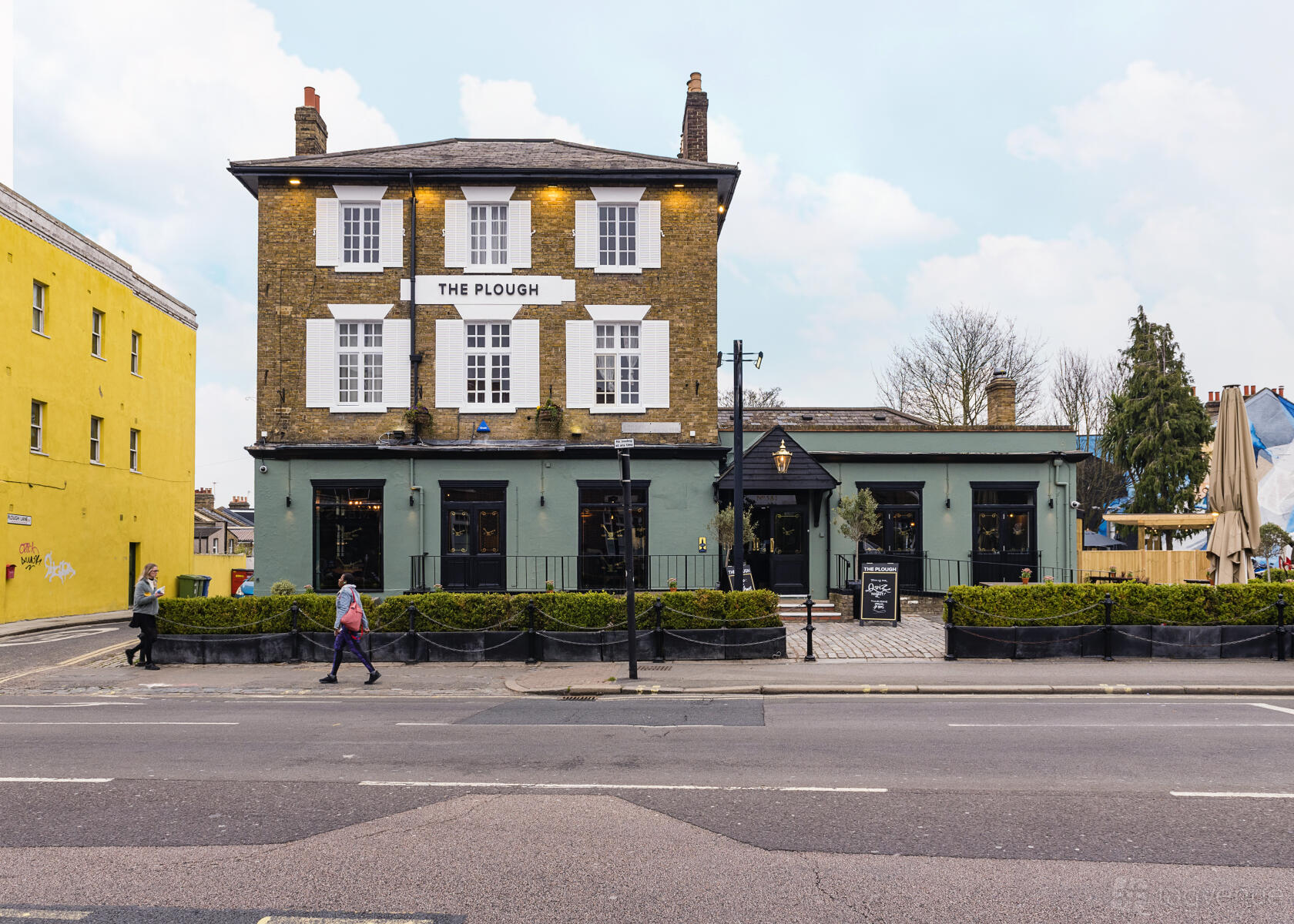 A traditional pub with exposed brick upper walls, large white-trimmed windows, and sage green facade at The Plough.