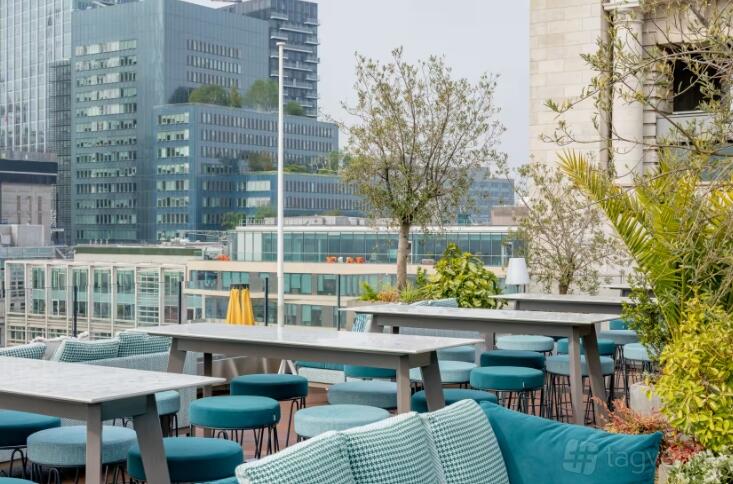 A rooftop bar with gray wooden tables, teal stools, and lush potted plants at Aviary Rooftop Bar & Restaurant.