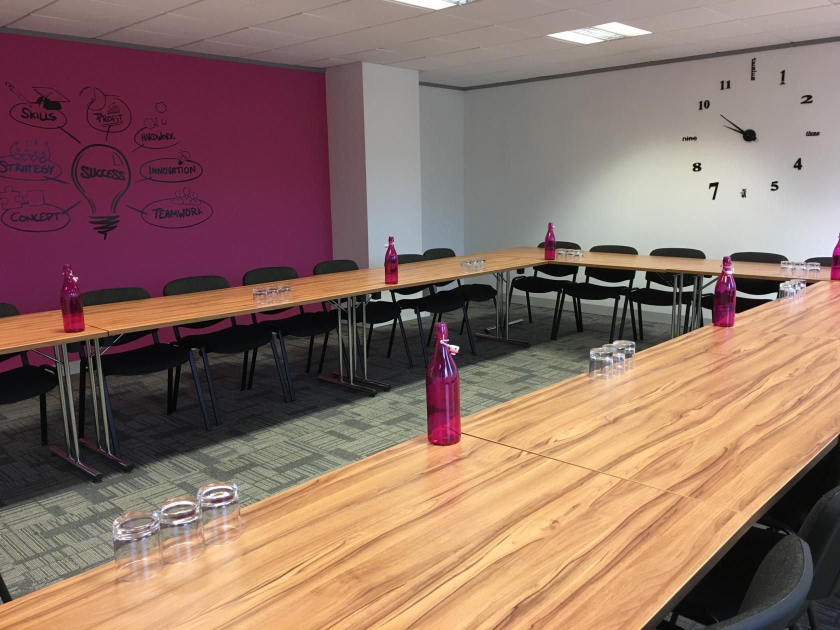 A meeting centre with U-shaped wooden tables, magenta water bottles, a large wall clock, and a pink feature wall at UBCUK Warrington at Rutherford House.