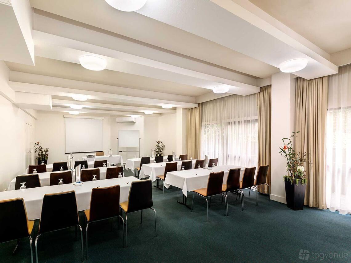 A hotel meeting room with rows of tables draped in white linens, floor-to-ceiling windows, and potted plants at Best Western Plus Travel Inn Hotel.