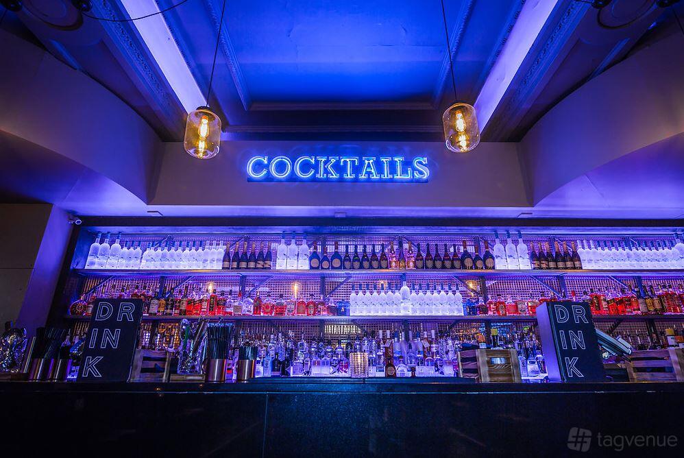 A bar with neon cocktail signage, glass shelves with illuminated bottles, and pendant lighting at Sway.