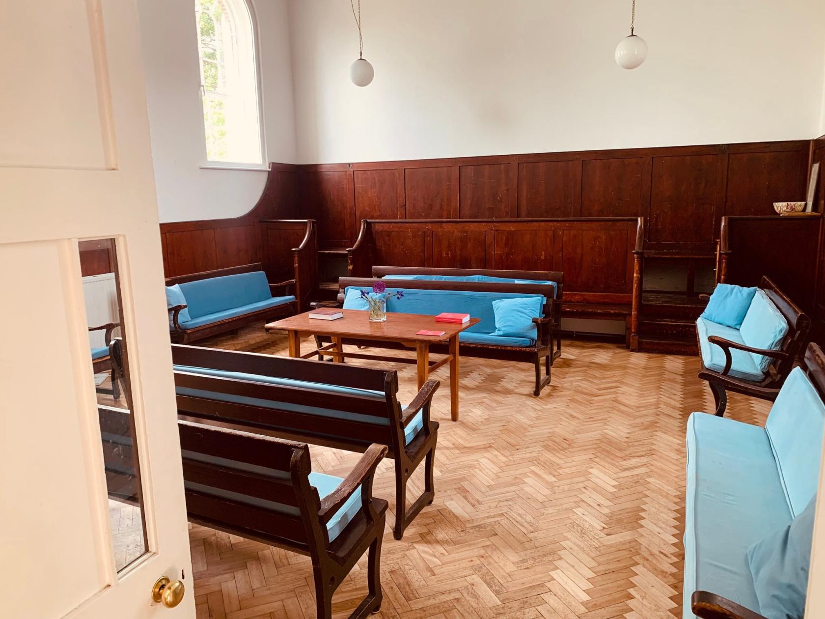 A church meeting room with wooden benches, blue cushions, parquet flooring, and a central table at Brentford & Isleworth Quaker Meeting House.