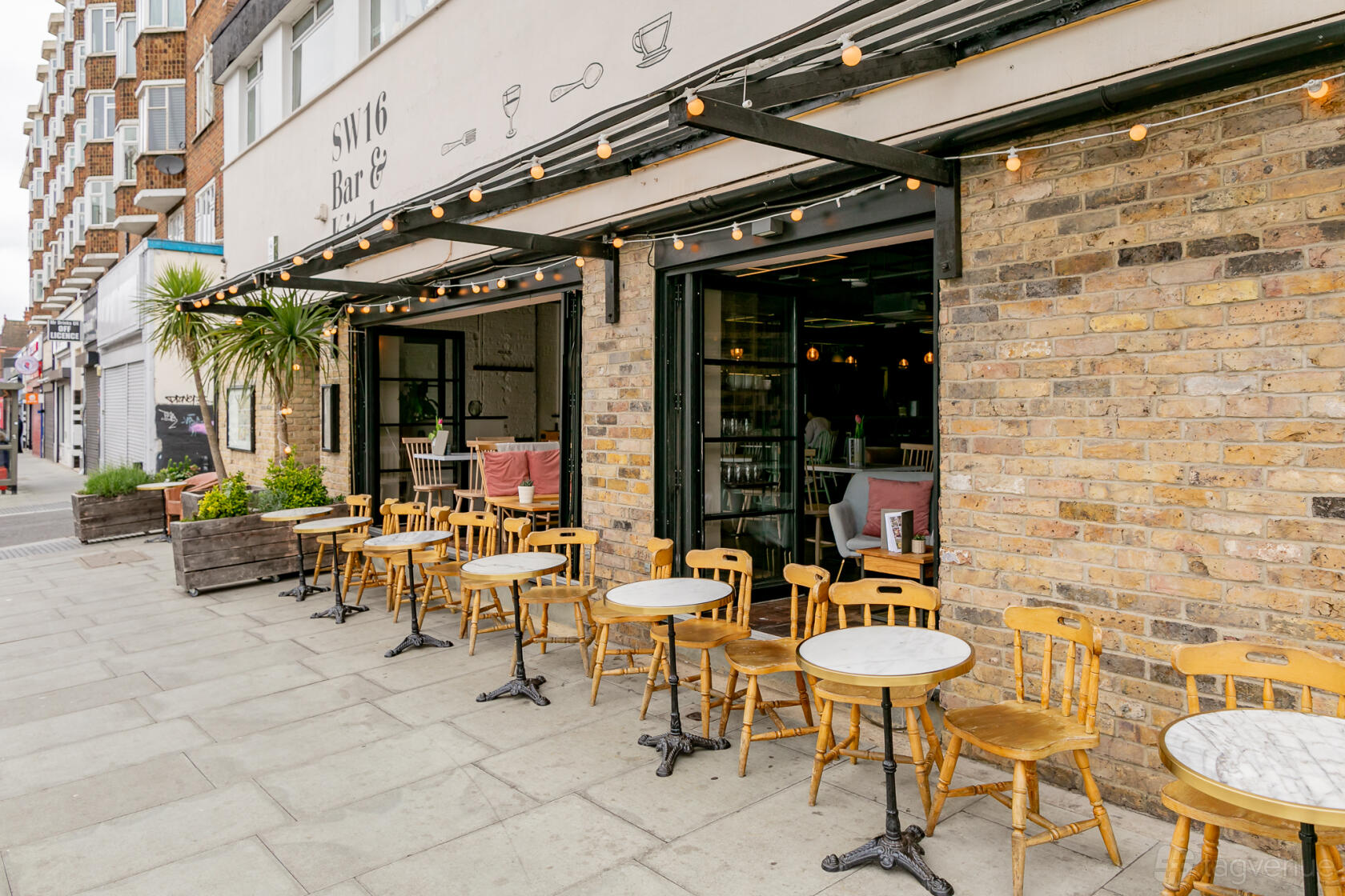 An outdoor restaurant area with round marble tables, wooden chairs, and exposed brick facade at SW16 Bar & Kitchen.