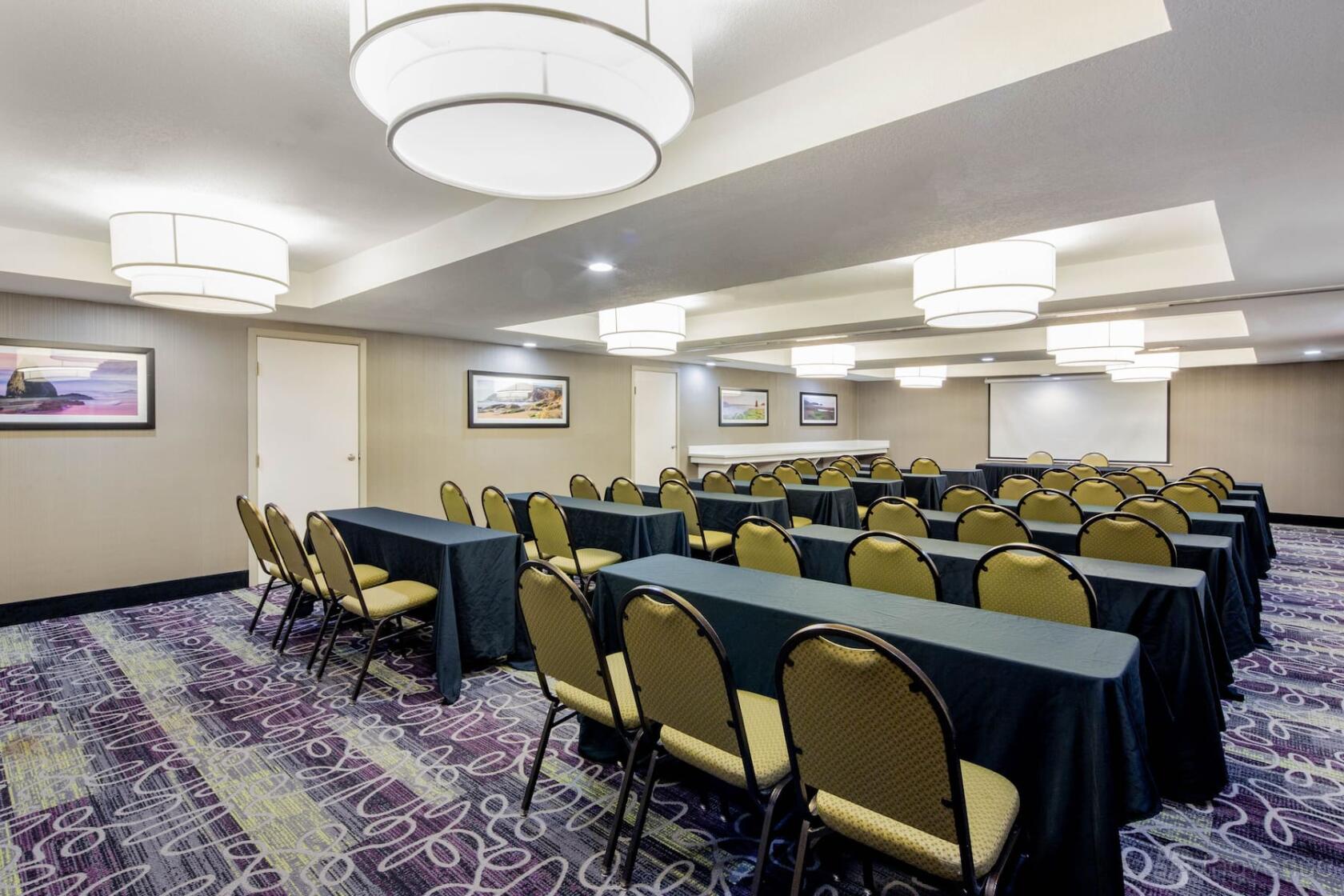 A hotel hall with rows of black-draped tables, cushioned chairs, and modern pendant lighting at La Quinta Inn & Suites by Wyndham.