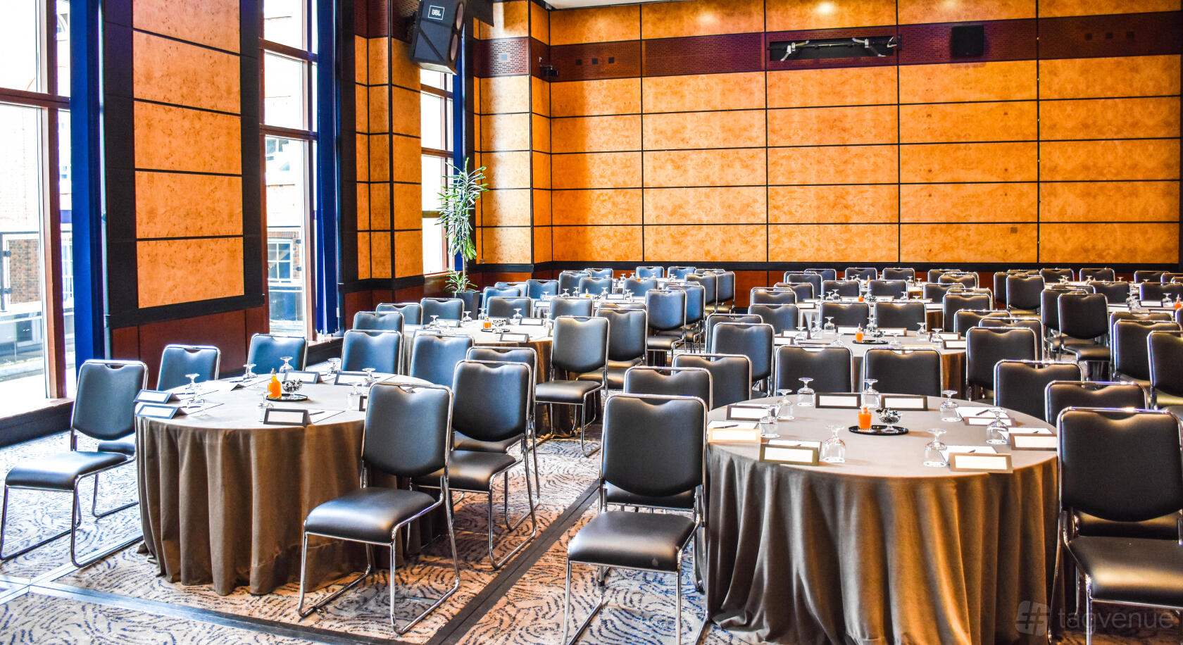 A hotel conference room with round tables, black chairs, wood-paneled walls, and floor-to-ceiling windows at Leonardo Royal London City.