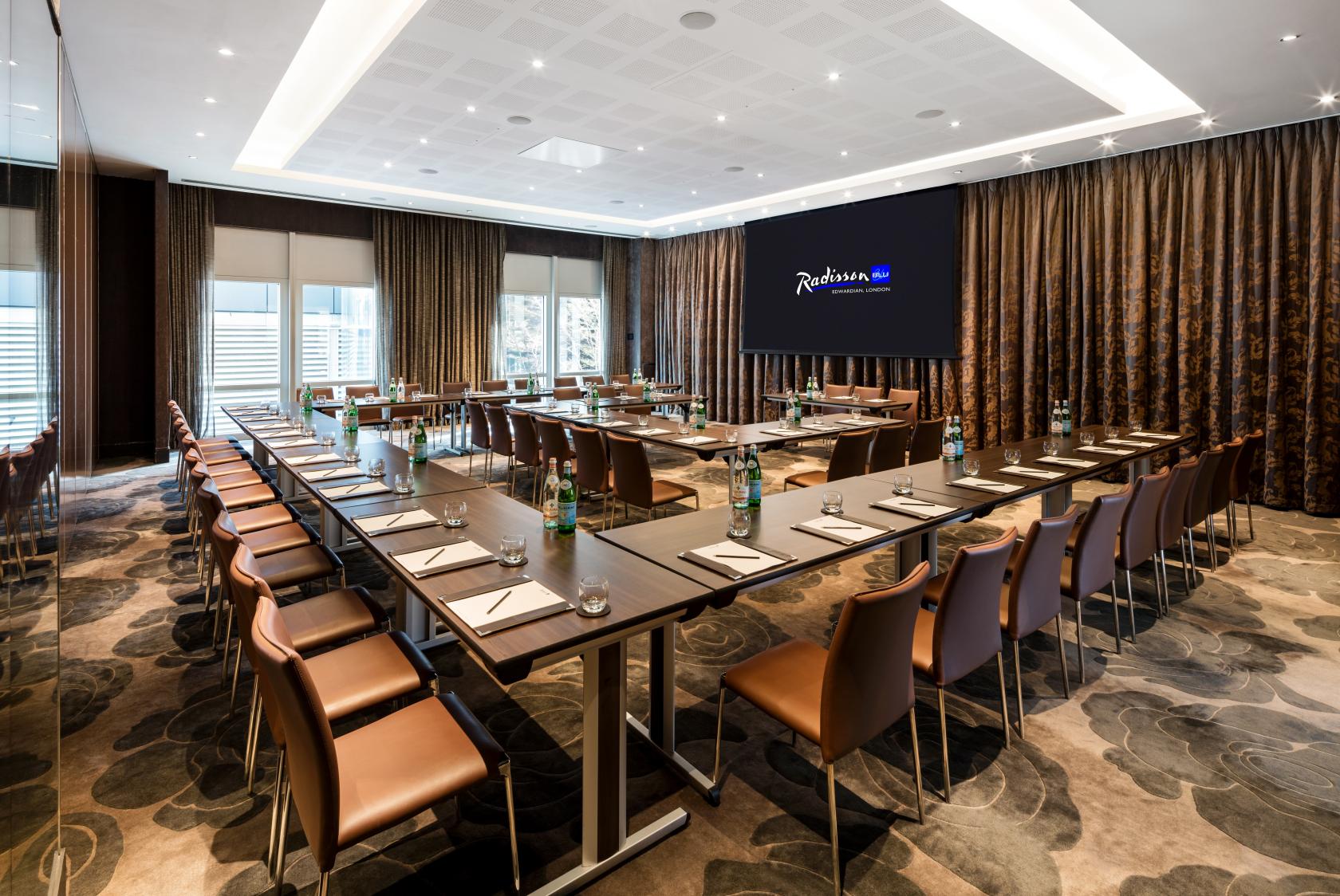 A meeting room with U-shaped tables, leather chairs, floor-to-ceiling windows, and a large presentation screen at Radisson Blu Hotel London Canary Wharf East.