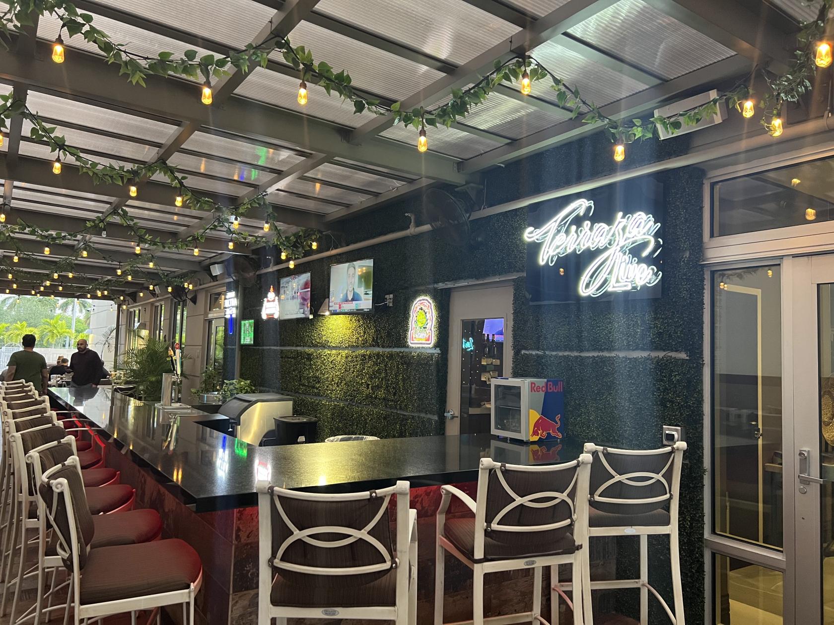 A covered terrace with a long bar, high chairs, string lights, and a green wall with neon signage at Tabbouleh Grill.