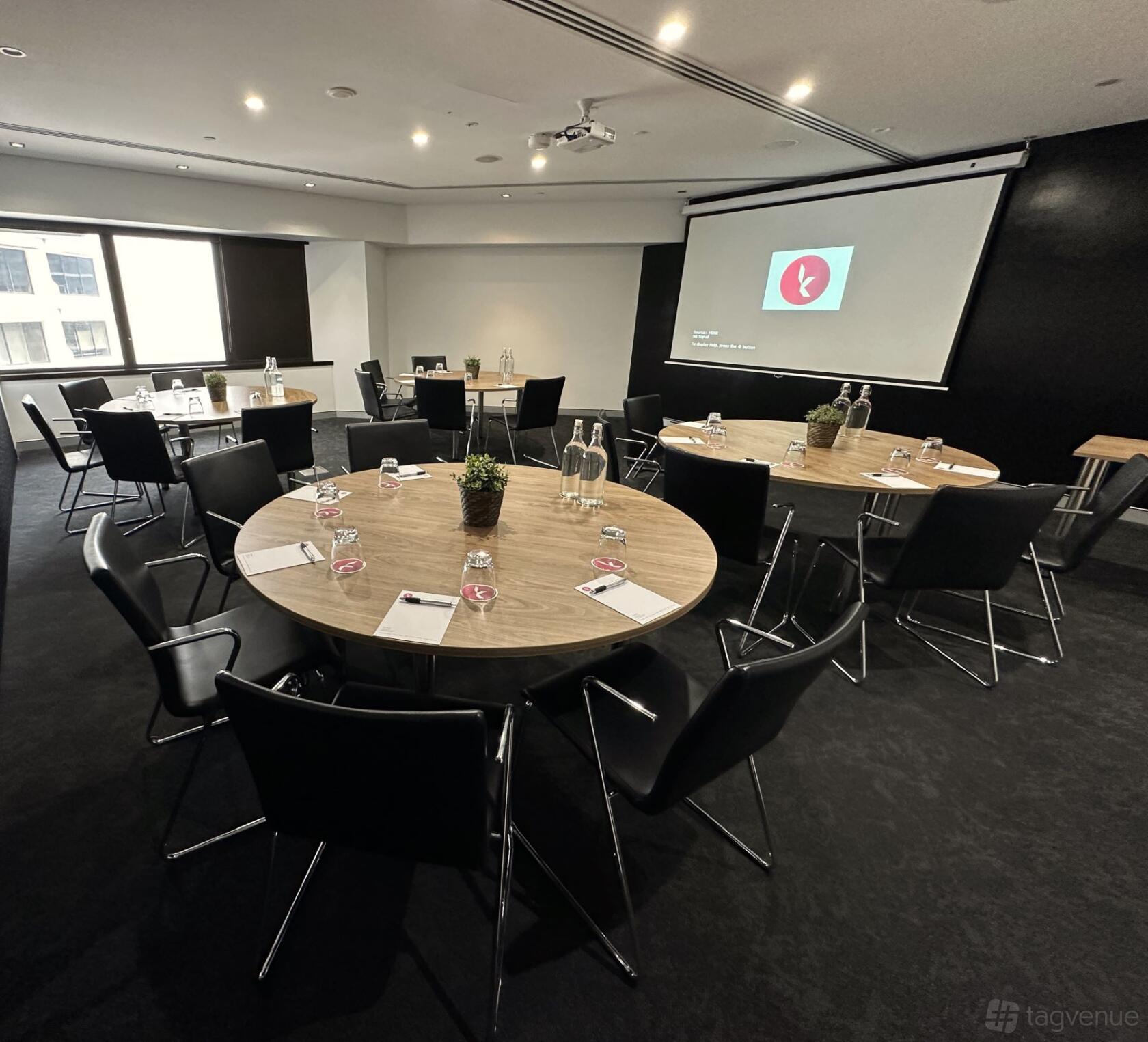 A conference centre with round tables, black chairs, a projector screen, and large windows at Karstens Brisbane.