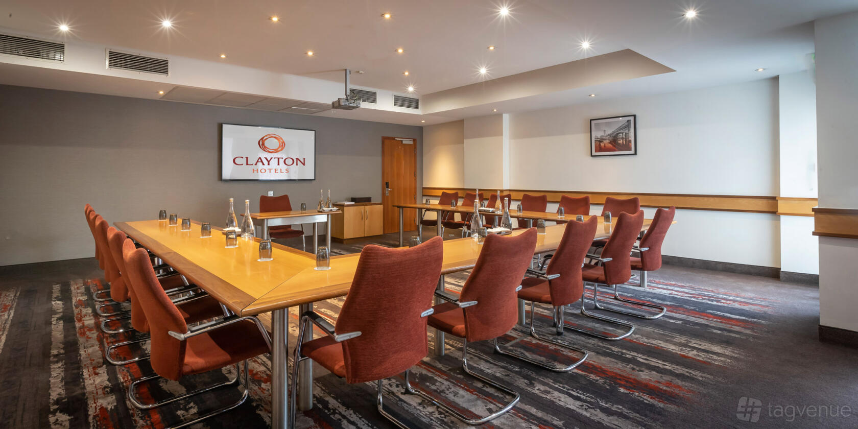 A hotel meeting room with U-shaped wooden tables, red chairs, and a projector screen at Clayton Hotel Manchester Airport.