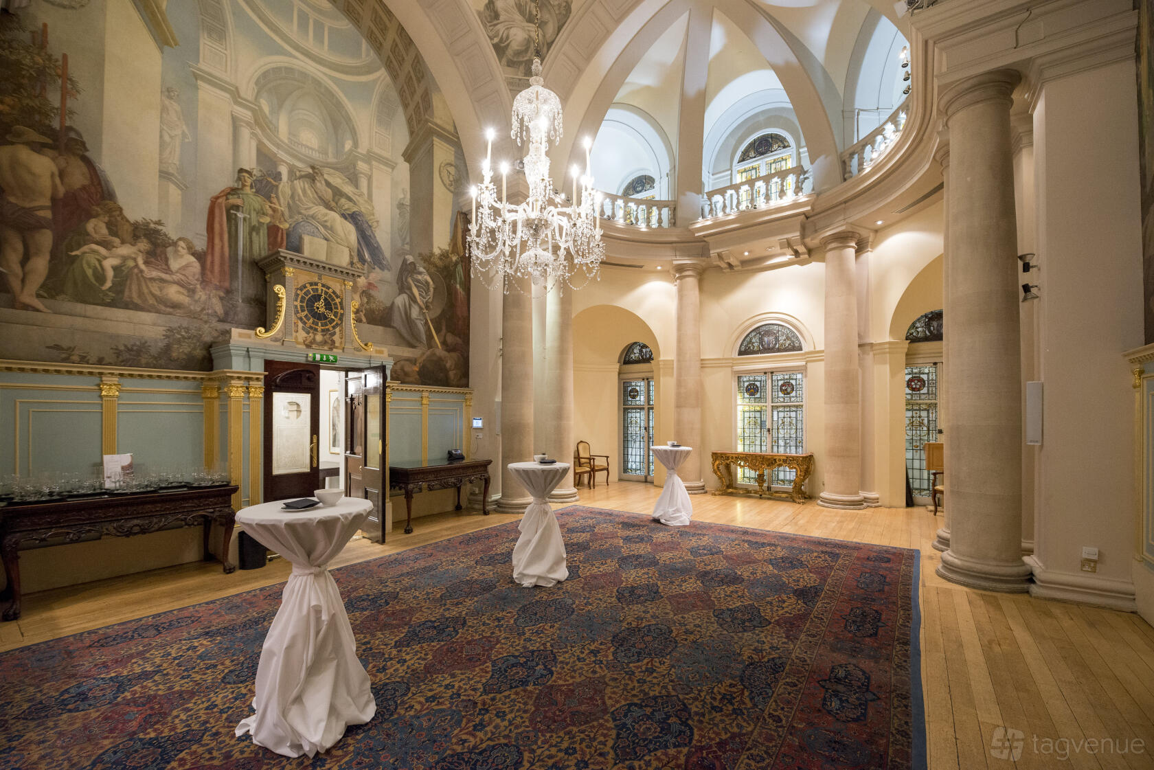 A grand hall with high arched ceilings, ornate chandeliers, wall murals, and tall windows at One Moorgate Place.